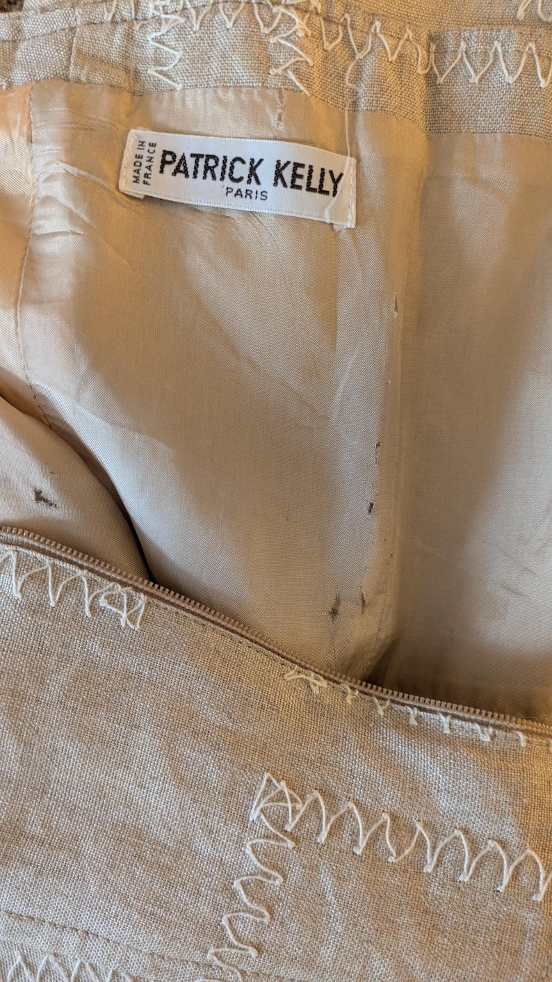 May include: A beige linen garment with a label that reads "Patrick Kelly Paris" and "Made in France". The garment has decorative stitching around the edges.