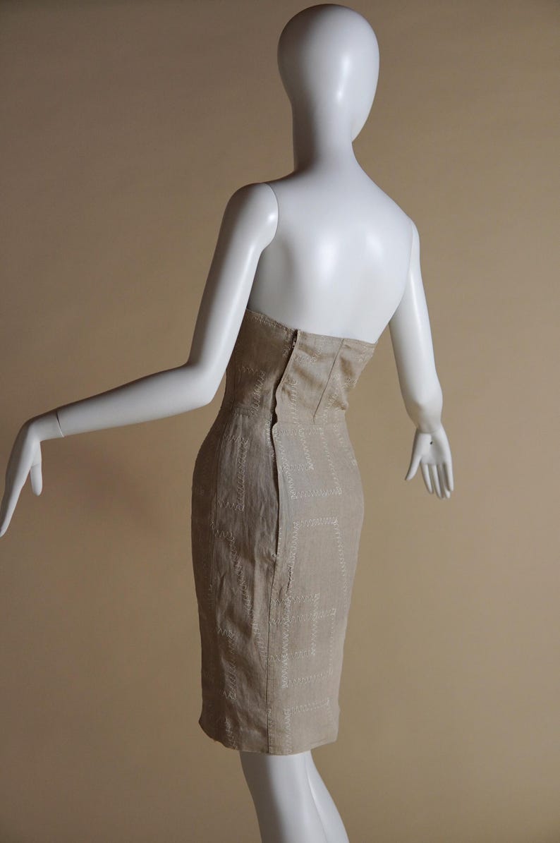May include: A beige strapless dress with a fitted silhouette and a geometric pattern. The dress is made of a textured fabric and has a zip closure at the back.