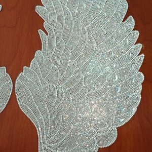 Puede incluir: Dos alas de ángel decorativas y con cuentas. Las alas son plateadas y blancas, con un patrón detallado de pequeñas cuentas. Las alas son simétricas y parecen estar hechas de tela o un material similar. Las alas se muestran sobre una superficie de madera marrón.