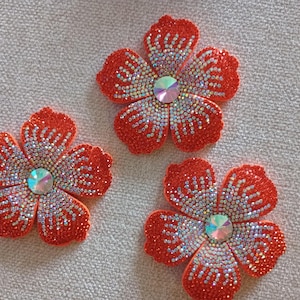 May include: Three five-petaled flower appliques in a vibrant orange hue. Each petal is densely covered in small, shimmering iridescent crystals that create a gradient effect. A large, circular, multifaceted clear crystal sits at the center.