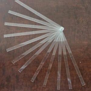 Könnte beinhalten: Ein fächerförmiges Zeichenwerkzeug aus klarem Acryl, bestehend aus mehreren dünnen, transparenten Streifen, die an einem einzigen Drehpunkt verbunden sind. Jeder Streifen weist kleine, gleichmäßig verteilte Löcher auf, die vom zentralen Metallverschluss ausgehen.