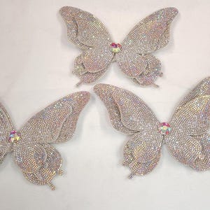 Könnte beinhalten: Drei glitzernde Schmetterlingsverzierungen. Jeder Schmetterling ist mit schillernden Strasssteinen bedeckt, mit einem zentralen Juwel. Die Flügel sind detailliert und ausgebreitet, wodurch ein schimmernder Effekt entsteht. Diese dekorativen Schmetterlinge sind ideal für Bastelarbeiten.