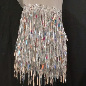May include: A white garment featuring a horizontal band of silver sequins at the top, from which hang numerous long, reflective silver fringe strips that shimmer with iridescent highlights, creating a dense, layered, and metallic appearance.
