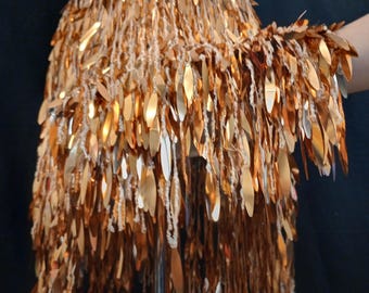 May include: A skirt featuring a dense layer of metallic gold, leaf-shaped sequins attached to a white crocheted base. The sequins are elongated and oval-shaped, creating a fringe-like effect that hangs vertically from the waistband down to the hem.