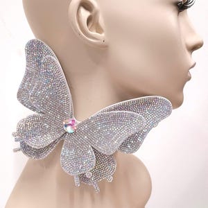 Può includere: Una scintillante collana a farfalla. La collana presenta grandi ali di farfalla a strati, ricoperte di strass scintillanti. Una gemma centrale iridescente accentua il design. La collana si indossa intorno al collo, creando un accessorio drammatico.