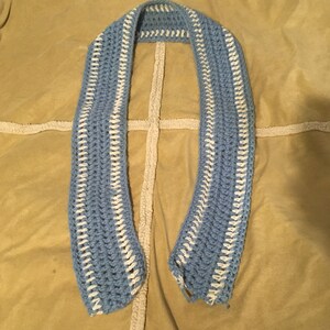 May include: A blue and white crocheted scarf with a white border. The scarf is laid out flat on a brown fabric surface.
