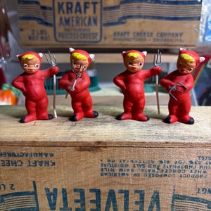 May include: Four small, vintage devil figurines. Each red figurine has a yellow head, white horns, and black feet. They hold small pitchforks. The figurines are displayed on a wooden box with text.