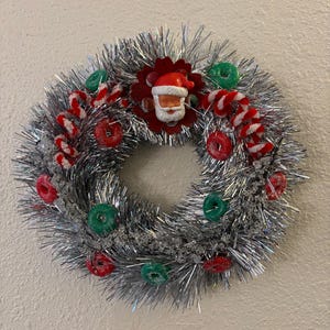 Pode incluir: Uma grinalda de Natal festiva feita de tinsel prateado, adornada com bengalas doces vermelhas e brancas, doces vermelhos e verdes e uma figura do Pai Natal. A grinalda circular é projetada para decoração de festas.