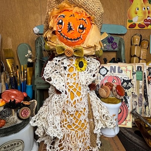 May include: A handcrafted scarecrow doll featuring a pumpkin head, straw hat, and a crocheted dress. The pumpkin face is painted with a smiling expression. A sunflower and a bow decorate the doll. The doll is displayed with other vintage items.