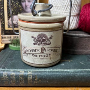 Pode incluir: Um pote de cerâmica bege com uma alça de arame metálico e uma tampa correspondente. A parte frontal apresenta um rótulo com o texto "LIMONADE PURGATIVE DE ROGE" e uma pequena ilustração de um sol sobre um livro aberto.