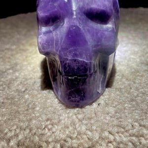 May include: A carved amethyst skull sculpture. The skull is a deep purple color with lighter streaks and variations in the stone. The details of the skull, including the eye sockets, nose, and teeth, are clearly defined. The sculpture is sitting on a textured surface.