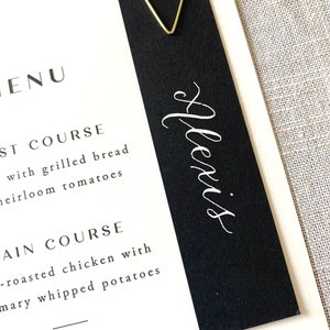 May include: A black and white menu card with a gold paper clip. The menu card has the text "MENU" at the top and lists the courses: "First Course" and "Main Course". The name "Alexis" is written in cursive on the black portion of the card.