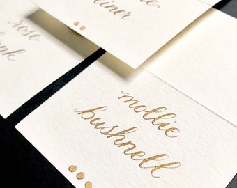 May include: Three white place cards with gold calligraphy. The names "Darlene Weiner", "Mollie Bushnell", and "Rose Pink" are written on the cards. Each card has three gold dots.