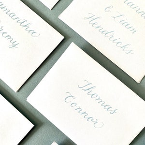 May include: Close-up of several white place cards with elegant, light blue calligraphy. Names like "Thomas Connor" and "Shanna & Liam Hendricks" are visible. The cards are arranged on a gray surface, suggesting a wedding or event theme.