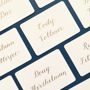 May include: A set of white cards with gold calligraphy names written on them. The cards are arranged on a dark blue surface.