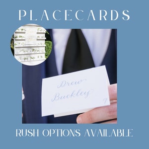 May include: A white place card with blue calligraphy that reads "Drew Buckley" is held by a person wearing a black tie and a blue suit jacket. The place card is part of a larger display of place cards on a white wall with greenery.