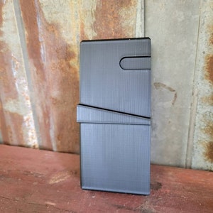 May include: A dark gray, rectangular wallet with a textured surface. The wallet features a curved cutout on the top and a diagonal line across the front. The wallet is standing upright on a red wooden surface.