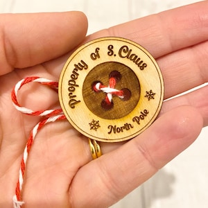 Puede incluir: Un adorno de botón de madera con el texto "Property of S. Claus North Pole" y una cuerda roja y blanca atada.