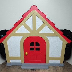 May include: A house-shaped Switch dock in red, beige, and white, with a red door and roof. The dock is designed to hold a Nintendo Switch console, with the controllers visible on either side.