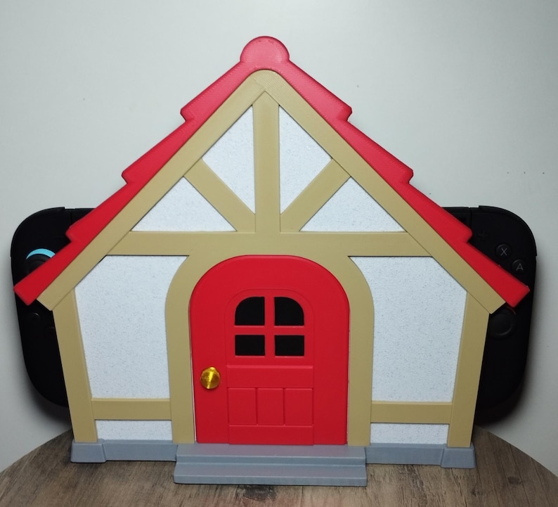 May include: A house-shaped Switch dock in red, beige, and white, with a red door and roof. The dock is designed to hold a Nintendo Switch console, with the controllers visible on either side.