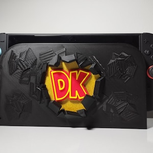 May include: A black Nintendo Switch console with a custom black dock featuring a 3D design of a wall being broken through. The words "DK" are in red with a yellow outline, set against a yellow background.