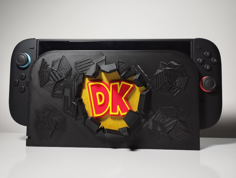 May include: A black Nintendo Switch console with a custom black dock featuring a 3D design of a wall being broken through. The words "DK" are in red with a yellow outline, set against a yellow background.
