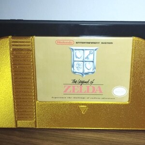 Może przedstawiać: Czarna przenośna konsola do gier umieszczona za złotym, teksturowanym plastikowym stojakiem w kształcie klasycznego kartridża do gry Nintendo Entertainment System. Etykieta zawiera napis The Legend of Zelda oraz ikonę tarczy.