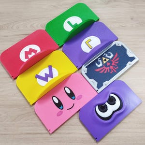 May include: A collection of colorful, stylized Nintendo-themed accessories. The items include a red "M" logo, a green "L", a yellow "W", a purple "L", a shield with a Triforce design, a pink Kirby face, and a purple Splatoon character.