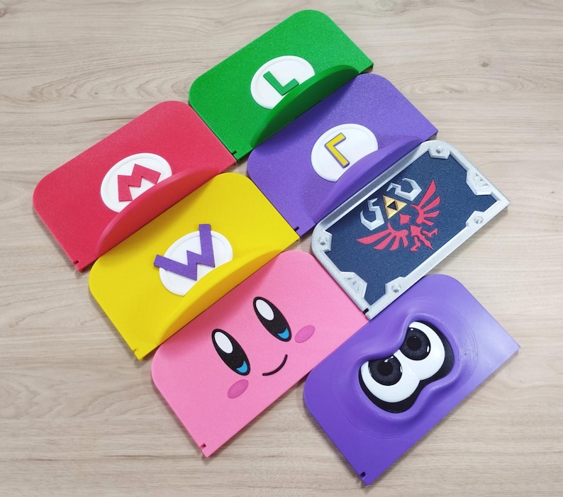 May include: A collection of colorful, stylized Nintendo-themed accessories. The items include a red "M" logo, a green "L", a yellow "W", a purple "L", a shield with a Triforce design, a pink Kirby face, and a purple Splatoon character.