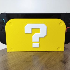May include: A black Nintendo Switch console rests on a bright yellow stand. The stand features a large white question mark, reminiscent of a classic video game. The stand has a rectangular shape with rounded corners.