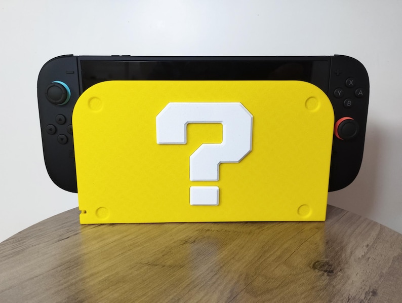 May include: A black Nintendo Switch console rests on a bright yellow stand. The stand features a large white question mark, reminiscent of a classic video game. The stand has a rectangular shape with rounded corners.