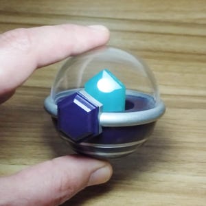 Pode incluir: Uma pequena cúpula de plástico transparente envolve um objeto em forma de cristal de cor azul turquesa. A cúpula fica sobre uma base roxa e prateada com uma gema hexagonal roxa na lateral.
