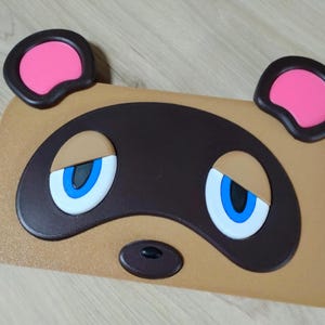 May include: A tan-colored, rectangular item featuring a cartoon face with large blue eyes, a black nose, and two pink-filled ears. The design is reminiscent of a popular video game character.
