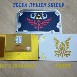 May include: Three decorative items inspired by the Zelda video game series. The top item is a blue and silver shield with a red emblem. Below is a gold NES cartridge and a white item with a gold emblem. Text includes "ZELDA HYLIEN SHIELD", "NES CARTRIDGE", and "RAURU".