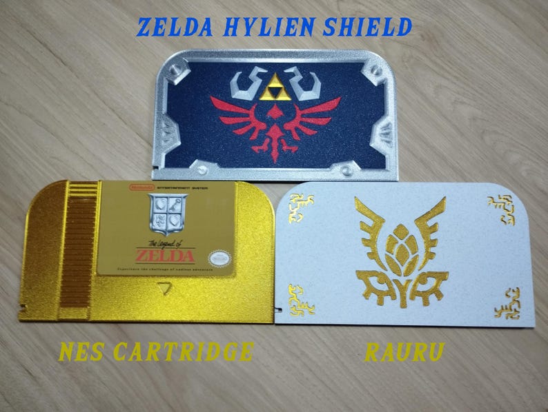 May include: Three decorative items inspired by the Zelda video game series. The top item is a blue and silver shield with a red emblem. Below is a gold NES cartridge and a white item with a gold emblem. Text includes "ZELDA HYLIEN SHIELD", "NES CARTRIDGE", and "RAURU".