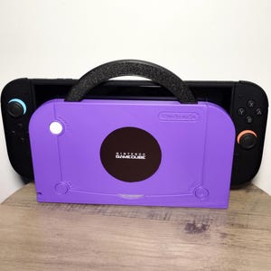 May include: A purple Nintendo GameCube-themed carrying case with a black handle, holding a black Nintendo Switch console. The GameCube case has a circular logo and the words "Nintendo GameCube" printed on it. The Switch has blue and orange buttons.