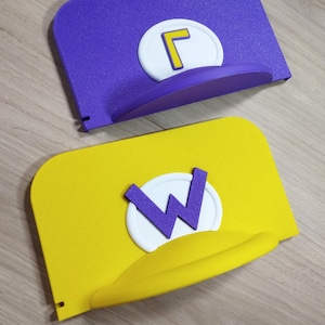 May include: Two novelty hats, one purple with a yellow "L" on a white circle, and one yellow with a purple "W" on a white circle. Both hats have a curved brim and are displayed on a wooden surface.