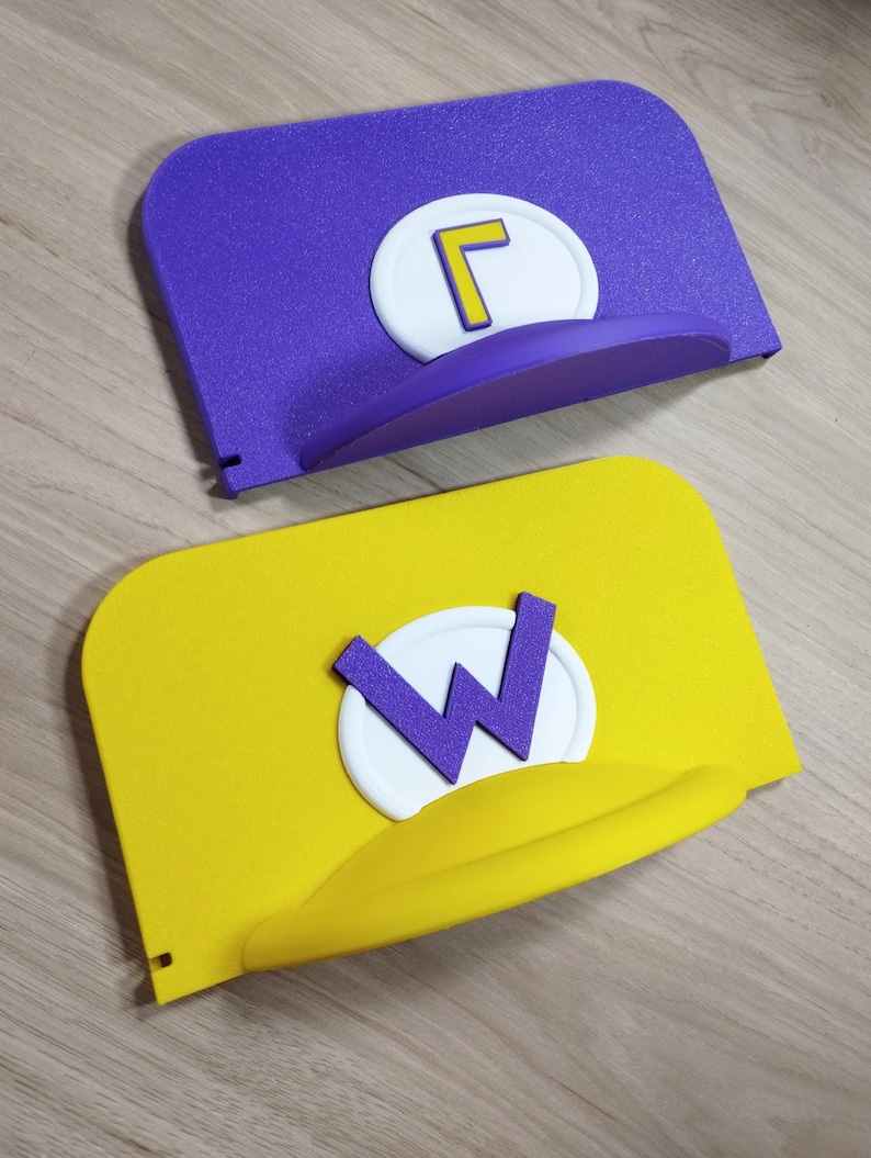 May include: Two novelty hats, one purple with a yellow "L" on a white circle, and one yellow with a purple "W" on a white circle. Both hats have a curved brim and are displayed on a wooden surface.