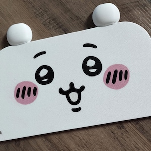 May include: A white, rectangular decorative panel with a cartoon face design. The face features large black eyes, pink blush marks, and a smiling mouth. Two white, spherical shapes are attached to the top of the panel, resembling ears.