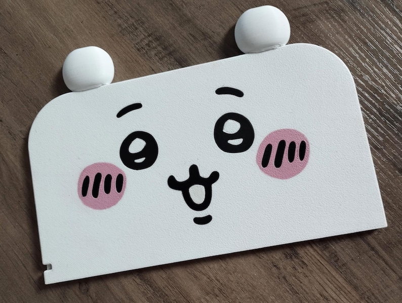 May include: A white, rectangular decorative panel with a cartoon face design. The face features large black eyes, pink blush marks, and a smiling mouth. Two white, spherical shapes are attached to the top of the panel, resembling ears.
