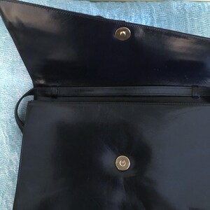 May include: Open view of a black leather handbag with a gold-colored clasp. The bag has a long strap and a rectangular shape. The interior is dark, and the leather has a glossy finish.