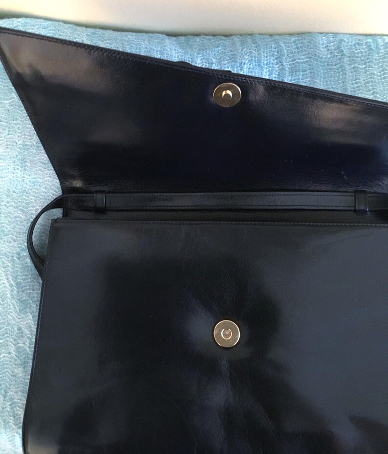 May include: Open view of a black leather handbag with a gold-colored clasp. The bag has a long strap and a rectangular shape. The interior is dark, and the leather has a glossy finish.