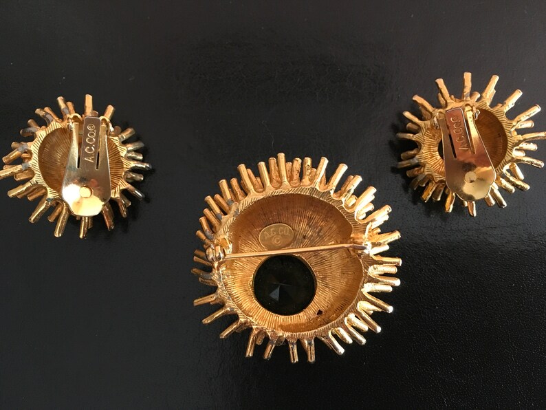 Pu&ograve; includere: Un set di tre gioielli dorati con un design a raggiera. La spilla ha una pietra nera al centro e gli orecchini sono rotondi. Il retro della spilla ha l'iscrizione "ACC 00", e gli orecchini hanno l'iscrizione "ACC 00".