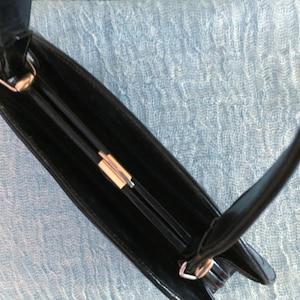 May include: A black handbag with a rectangular shape and a handle. The bag features a gold-colored clasp and a sleek, glossy finish. The interior of the bag is visible, revealing a dark compartment.