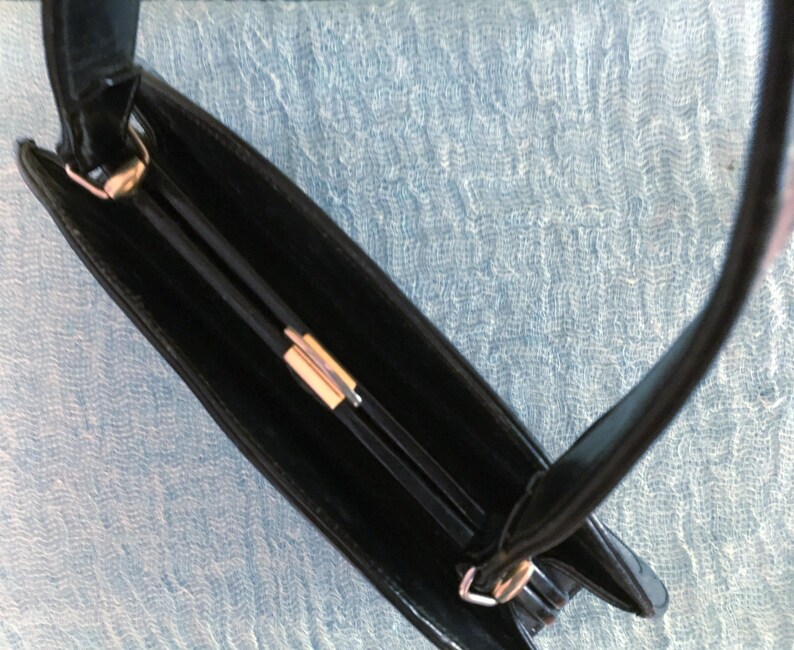 May include: A black handbag with a rectangular shape and a handle. The bag features a gold-colored clasp and a sleek, glossy finish. The interior of the bag is visible, revealing a dark compartment.