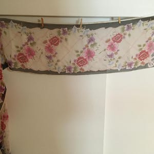 May include: A sheer white scarf with a floral print in shades of pink, purple, and green. The scarf has a dark grey border and is hanging on a clothesline.
