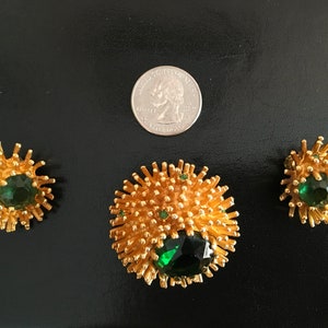Pu&ograve; includere: Un set di tre gioielli dorati con pietre preziose verdi. Il set comprende una spilla e un paio di orecchini. La spilla &egrave; pi&ugrave; grande degli orecchini e presenta una grande pietra preziosa verde al centro. Gli orecchini hanno pietre preziose verdi pi&ugrave; piccole al centro.
