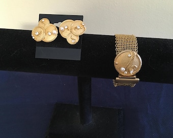 May include: A gold tone bracelet with a mesh chain and a large circular pendant featuring a pearl design. A pair of gold tone earrings with a similar pearl design are also shown.