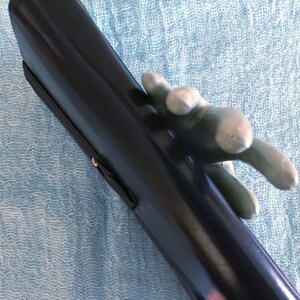 May include: A black, glossy, cylindrical object with a silver-colored hand-shaped handle. The object has a black strap attached to it. The background is a light blue, textured fabric.
