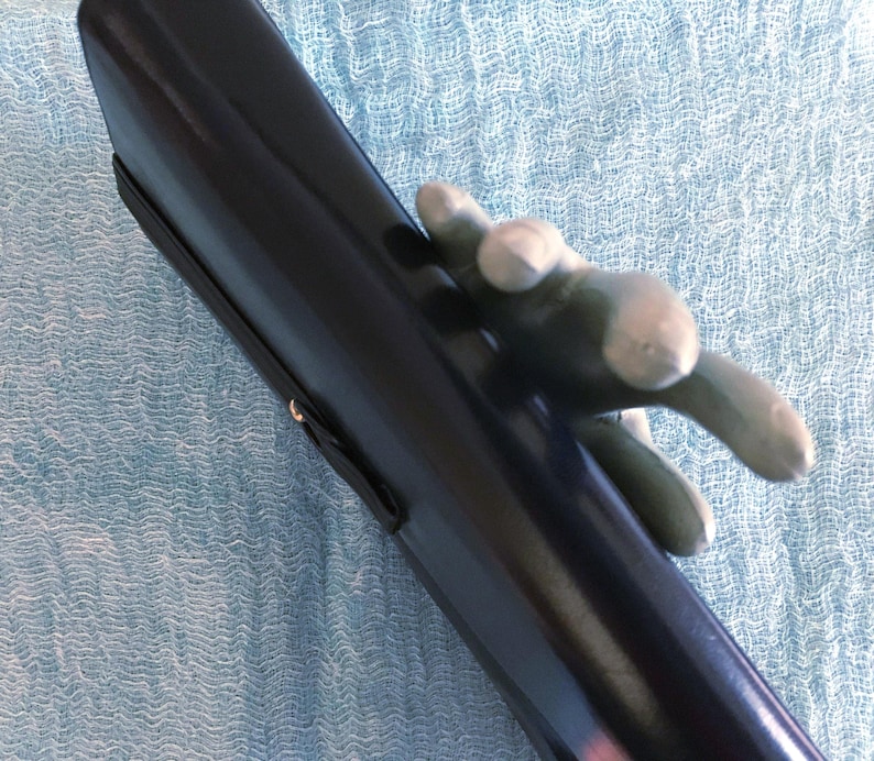 May include: A black, glossy, cylindrical object with a silver-colored hand-shaped handle. The object has a black strap attached to it. The background is a light blue, textured fabric.